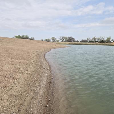 View of a gently sloped sandy shoreline curving around a calm off-channel basin, with trees and cloudy sky in the background at Cottonwood Ranch in Nebraska.
